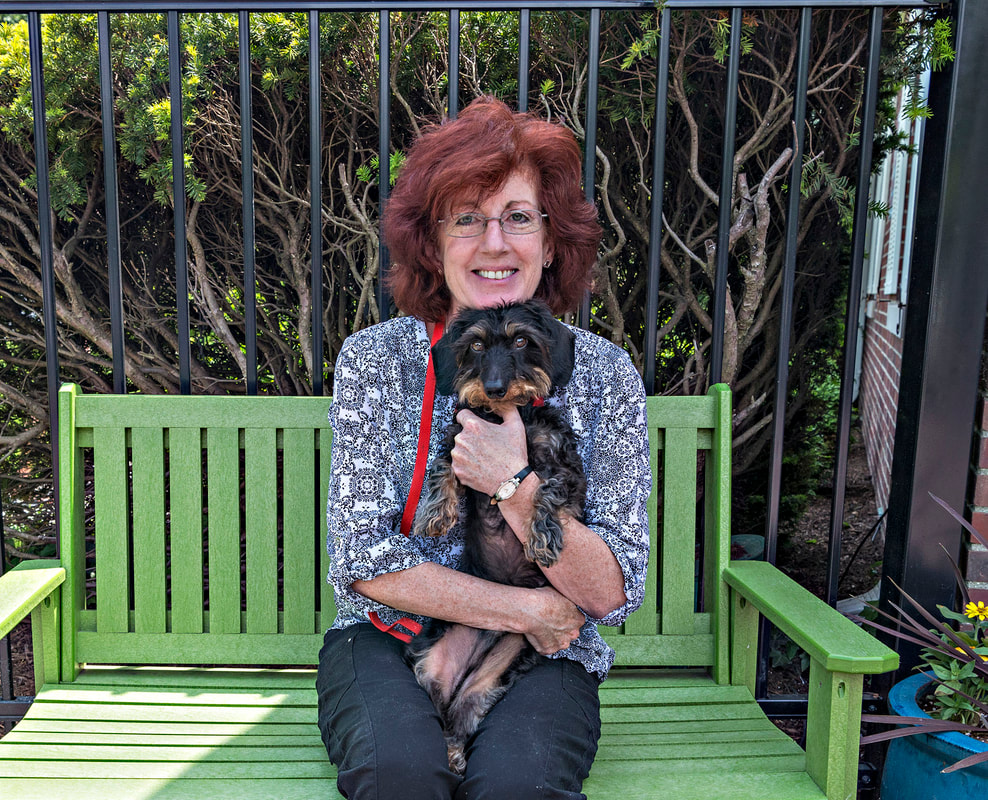 Photo of a woman with red hair holding a black and brown long haired Dachshund breed dog sitting on a green bench