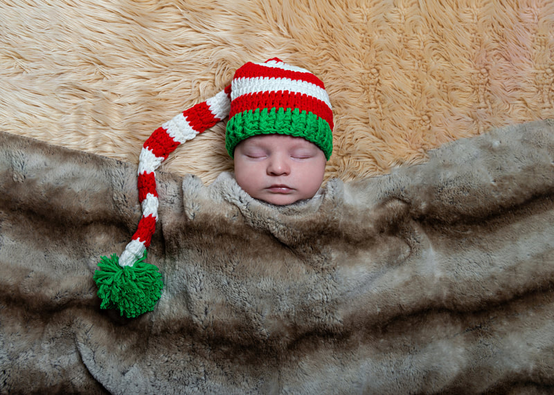 photo of a newborn baby boy laying on a beige furry rug under a faux chinchilla fur blanket wearing a white red and green long knit hat