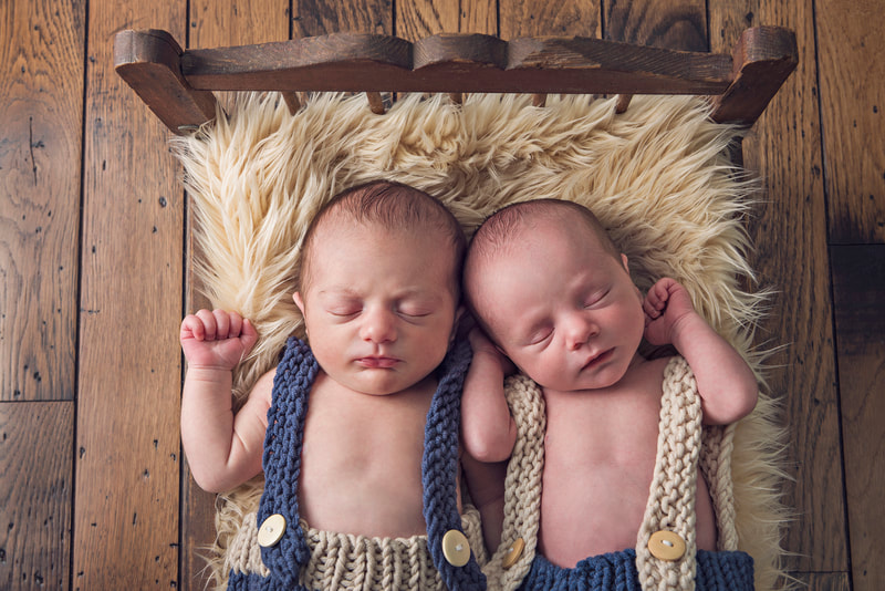 photo of newborn baby twin boys sleeping on a beige furry rug on a vintage doll bed against a brown hardwood floor. They are wearing blue and cream coordinated suspender outfits.