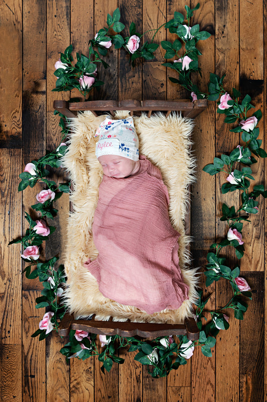 photo of a newborn baby girl in a pink cheesecloth wrap and an embroidered name hat laying on a beige furry rug on a vintage doll bed. She is against a hardwood floor and is surrounded by a garland of roses.