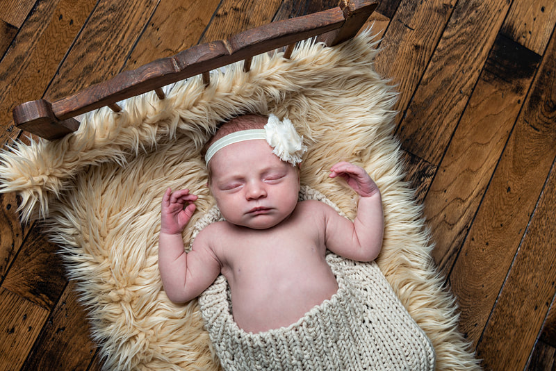 photo of a newborn baby girl in a beige knit wrap laying on a beige furry rug on a vintage wooden doll bed against a hardwood floor. She wears a white flower headband