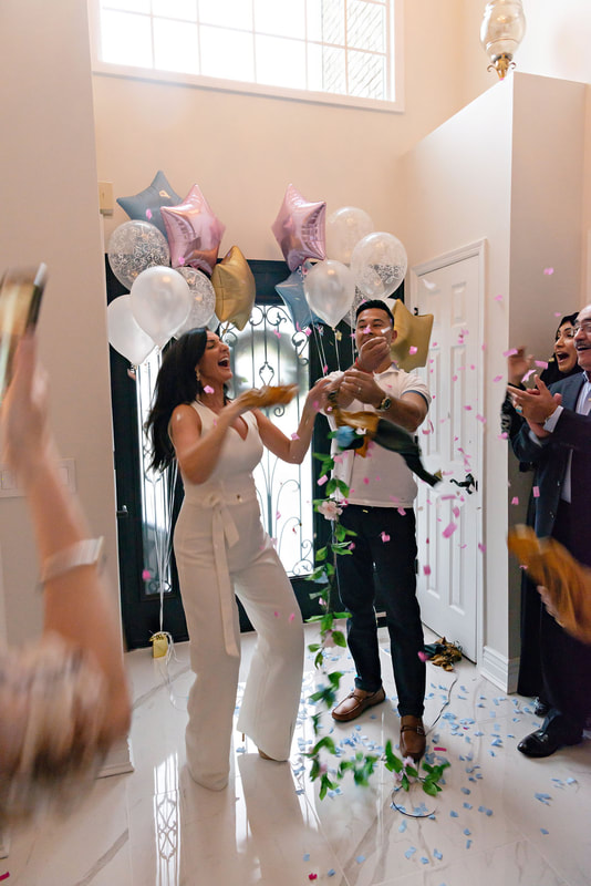 picture of a pregnant woman and her husband standing in an airy light-filled foyer in between bouquets of balloons popping a golden balloon filled with pink confetti, revealing the gender of their upcoming baby. 