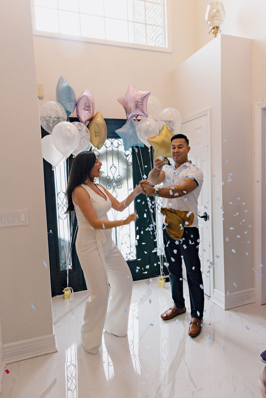 picture of a pregnant woman and her husband standing in an airy light-filled foyer in between bouquets of balloons popping a golden balloon filled with blue confetti, revealing the gender of their upcoming baby