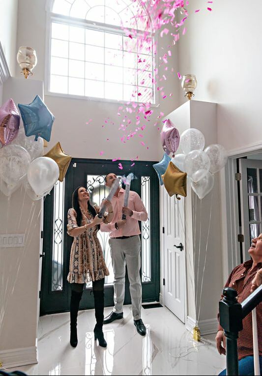 picture of a pregnant woman and her husband standing in an airy light-filled foyer in between bouquets of balloons shooting off a confetti cannon filled with pink confetti, revealing the gender of their upcoming baby. 