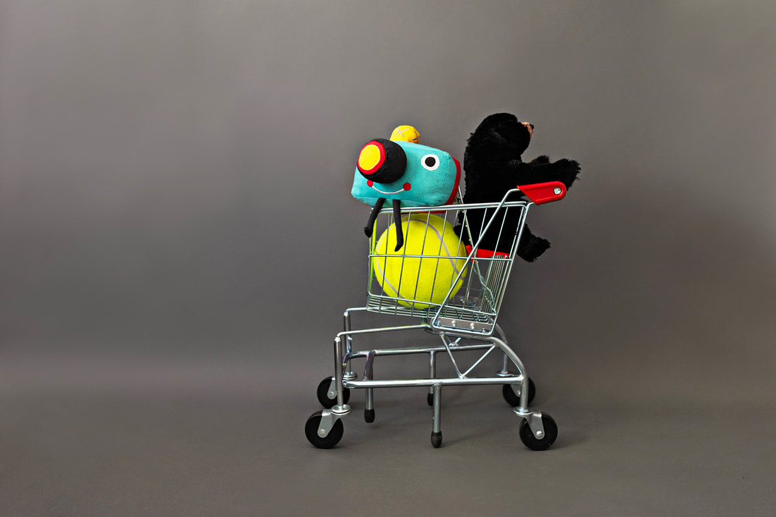 photo of a kids' toy shopping cart loaded with a stuffed bear, stuffed camera and an oversized tennis ball toy on a gray background