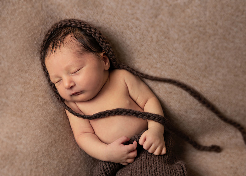 photo of a newborn baby boy wearing a brown knit hat and pants laying on a beige blanket