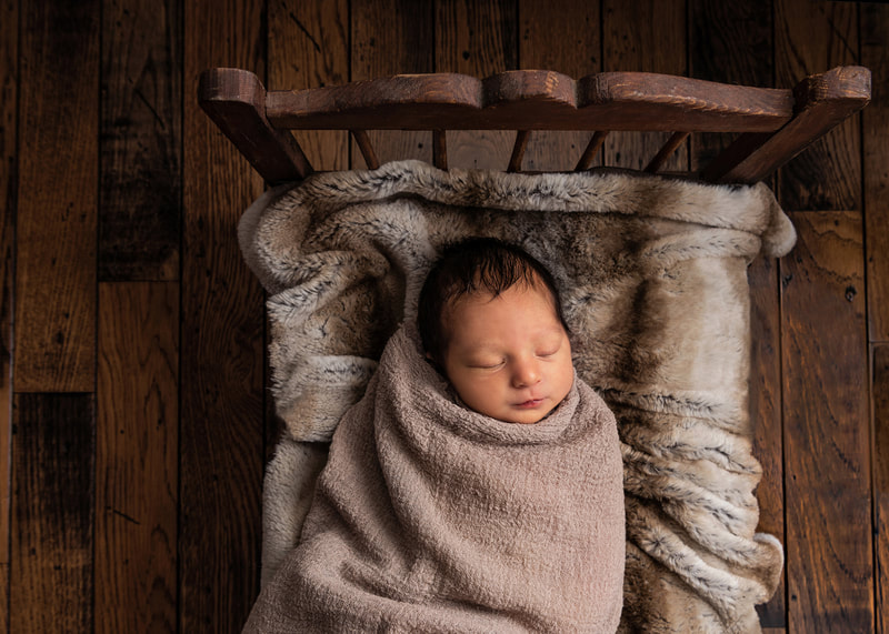 photo of a newborn baby boy wrapped in a beige wrap laying on a faux chinchilla blanket on a vintage doll bed against a hardwood floor in a photo studio