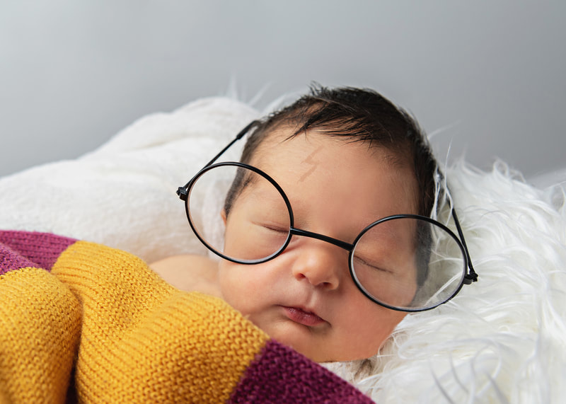 photo of a newborn baby boy laying on a white furry rug covered in a Harry Potter Gryffindor scarf, wearing Harry Potter glasses and  with a faint Photoshopped lightning bolt shaped scar on his forehead