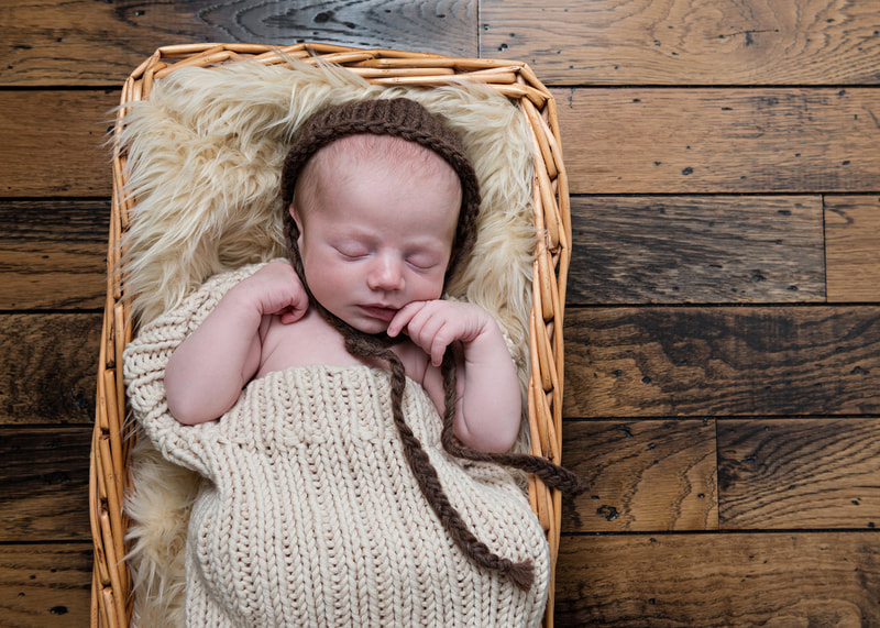 photo of a newborn baby boy wearing a beige knit wrap and a brown knit cap laying on a beige furry rug in a wicker basket against a brown hardwood floor