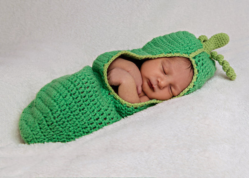 photo of a newborn baby boy laying on a white blanket dressed in a knit pea pod costume
