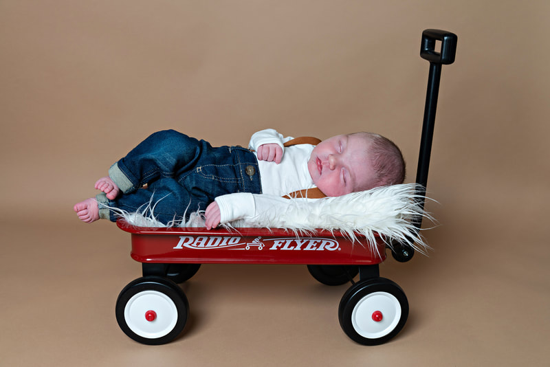 photo of a newborn baby boy in overalls and denim jeans laying on a furry white rug in a red Radio Flyer wagon against a beige paper backdrop in a photo studio