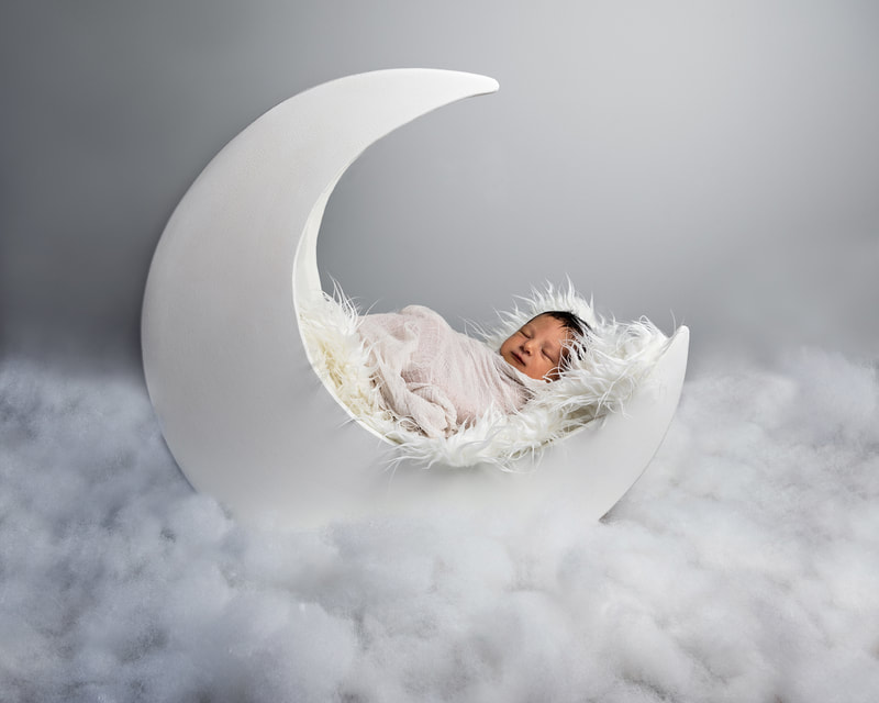 photo of a newborn baby boy wrapped in a white cheesecloth wrap laying on a white furry rug inside a white wooden half moon prop. He is surrounded by cotton on the ground resembling white clouds.
