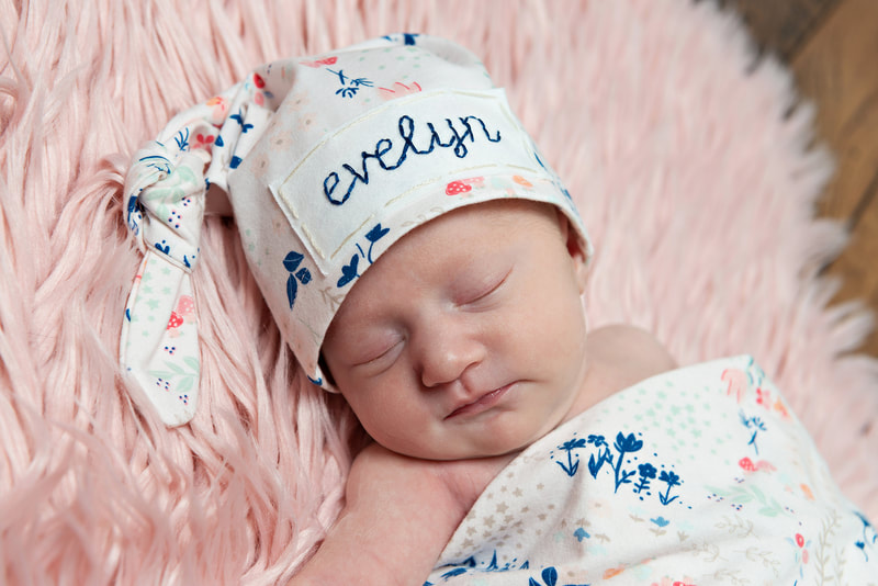 photo of a newborn baby girl wearing an embroidered hat with her name on it laying on a furry pink rug in a photo studio