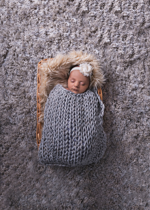 photo of a newborn baby girl laying on a beige furry rug in a wicker basket on top of a grey furry carpet. She wears a white flower headband and is covered with a grey knit blanket