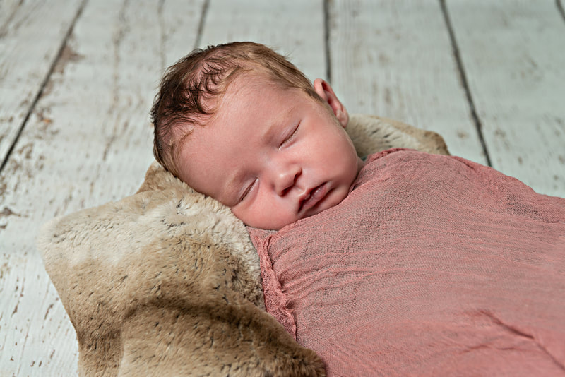 photo of a newborn baby girl wrapped in a pink cheesecloth wrap laying on a faux chinchilla fur rug against a white wooden backdrop