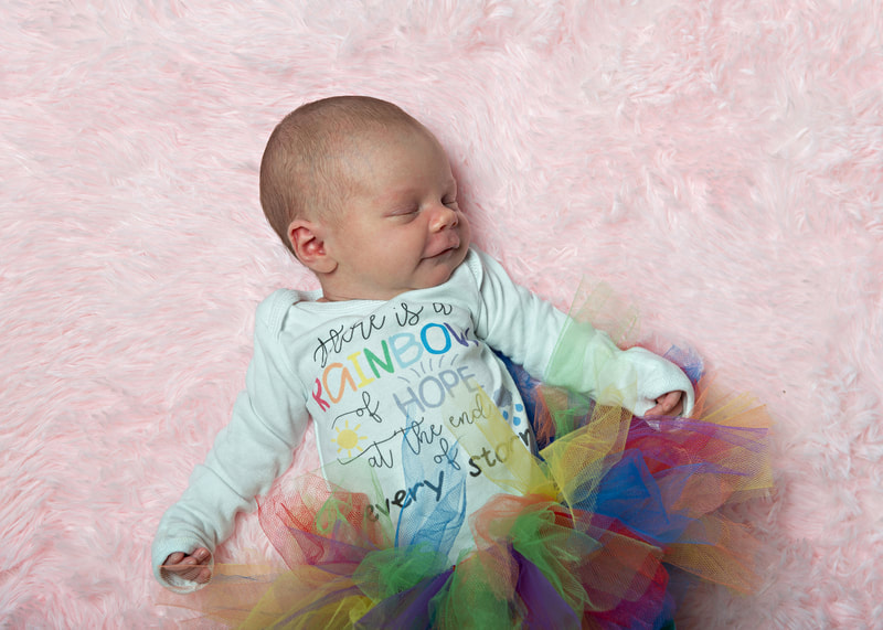 photo of a newborn baby girl wearing a rainbow tutu and a rainbow baby graphic onesie laying against a pink furry rug