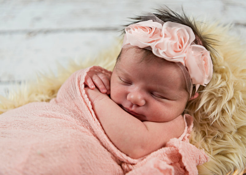 photo of a newborn baby girl wrapped in a pink cheesecloth wrap, wearing a pink flower headband and laying on a beige furry rug against a white wooden backdrop in a photo studio