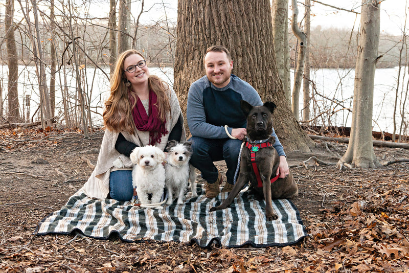 Photo of a woman and a man crouching and kneeling on a plaid blanket in the woods. They are posing with their two small white terrier mix dogs and their large black shepherd mix dog. 