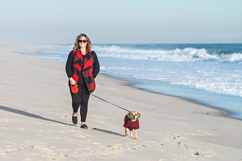 photo of a woman walking along a beach walking a puggle dog on a leash. They both wear matching red buffalo plaid outfirts. 